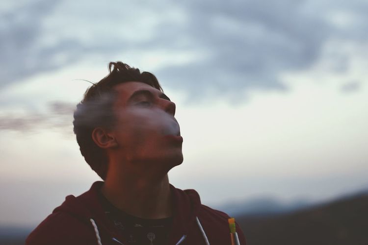 Young man smoking electronic cigarette against sky