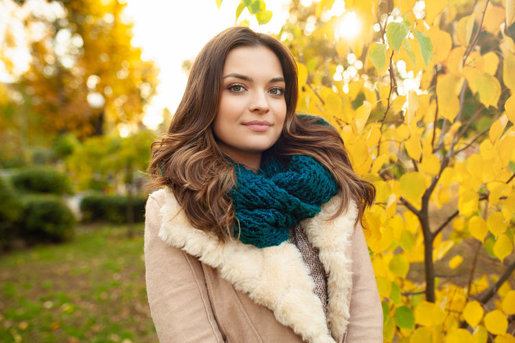 Portrait of beautiful young woman in warm clothing against autumn tree