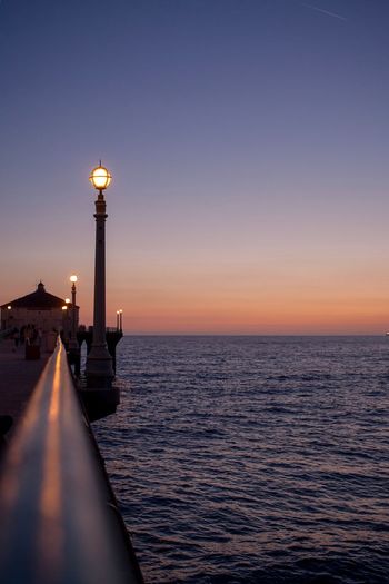 Illuminated street light by sea against sky during sunset