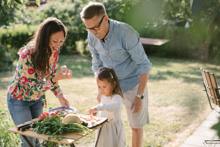 Girl cutting food while standing by mother and grandfather at table in backyard