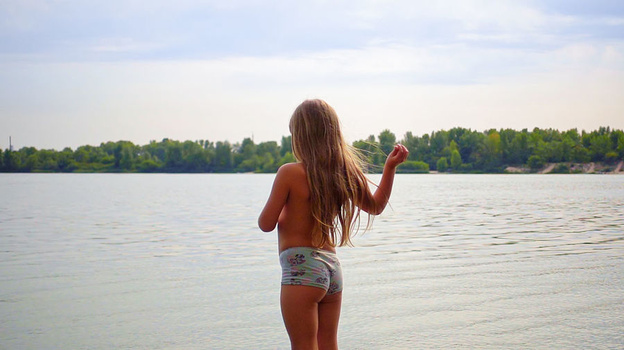 Shirtless girl standing at lakeshore against sky