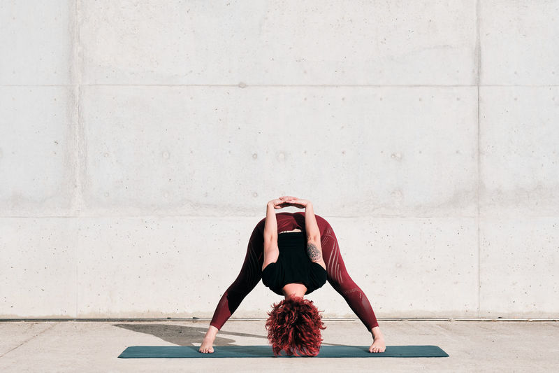 Trendy fit female athlete in sportswear doing wide legged forward bend yoga pose on sports mat while training alone on street against concrete wall in sunny day