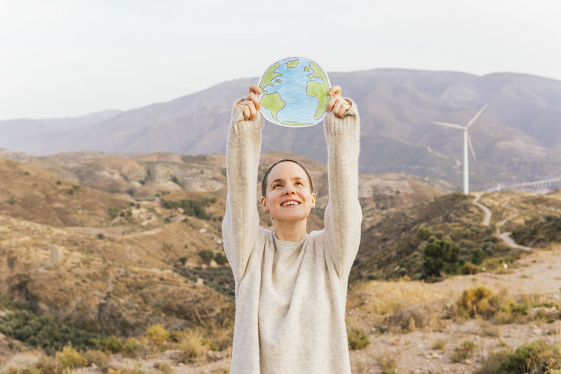 Smiling shaved head woman with hand raised holding planet earth on mountain