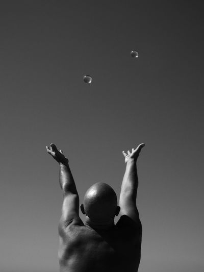 Rear view of bald man with arms raised towards bubbles against sky
