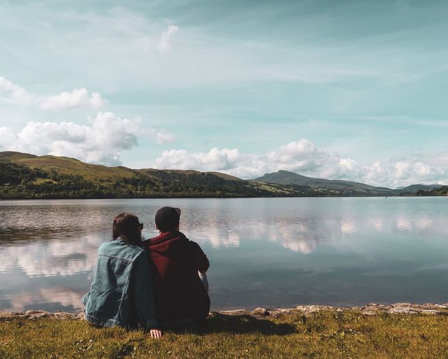 Rear view of couple sitting at lake against sky