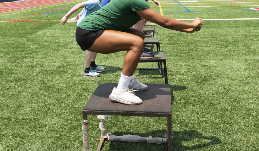 Side view of athletes jumping on different size plyo boxes outdoors on a green turf field.