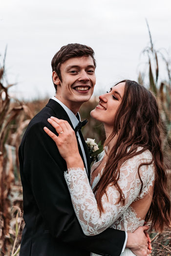 Smiling newlywed couple embracing while standing against sky