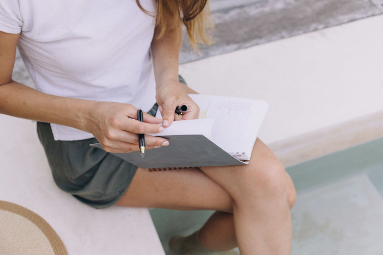 Midsection of woman with a notebook turning pages on her lap, dangling her legs on the pool