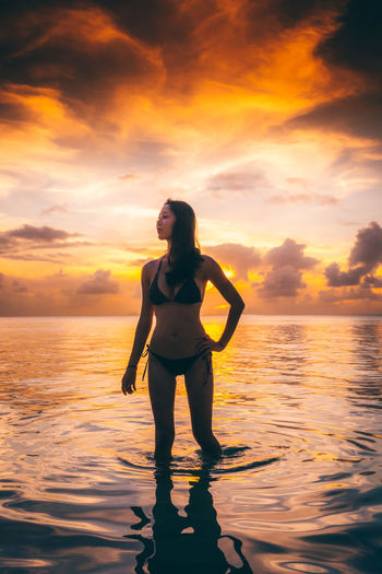 Woman standing in water at beach against orange sky during sunset