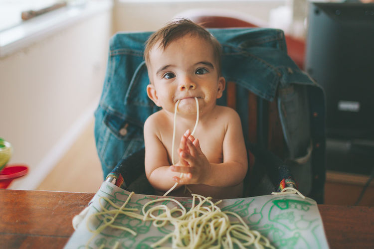 Portrait of shirtless baby boy eating spaghetti at home