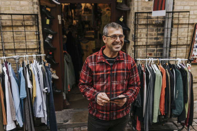 Happy male owner with mobile phone standing outside clothing store
