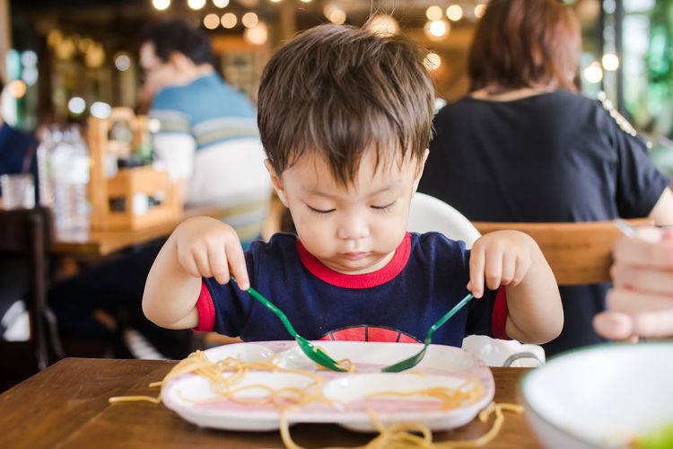 Cute baby boy playing with noodles at table in restaurant