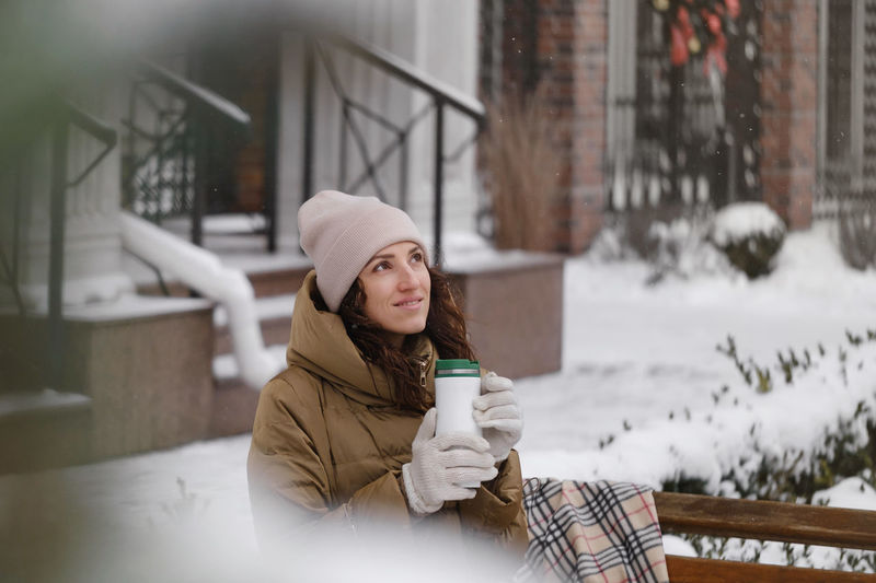 Cheerfully brunette woman drinking from flask outdoors in the city in winter. warming up, enjoying