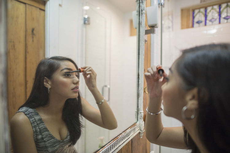 Young woman applying makeup in her bathroom mirror