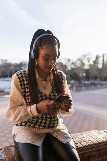Charming african american female with braids sitting on stone fence on plaza de espana and enjoying music in headphones while browsing smartphone
