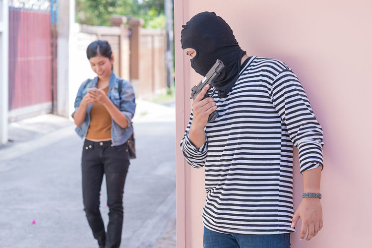 Thief with gun hiding against wall by smiling young woman walking on street by