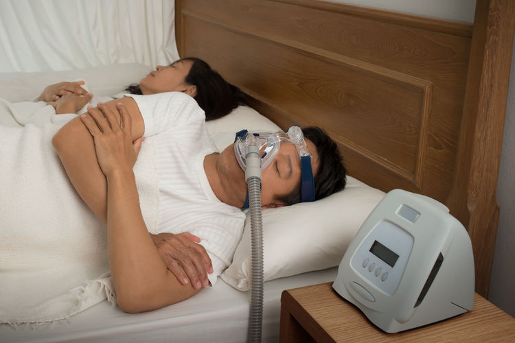 Man wearing cpap mask while suffering from sleep apnea by woman on bed at home