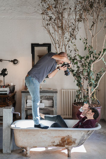 Side view dedicated man standing on bathtub edges and shooting from above adult guy in stylish clothes lying in aged white bath near big green potted tree against rustic vintage interior in photo studio