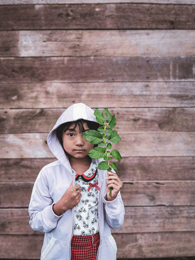 Portrait of girl holding plant standing against wooden wall outdoors