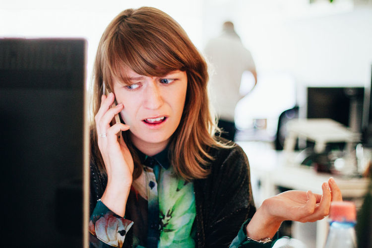 Worried woman talking on mobile phone while sitting at computer desk in office