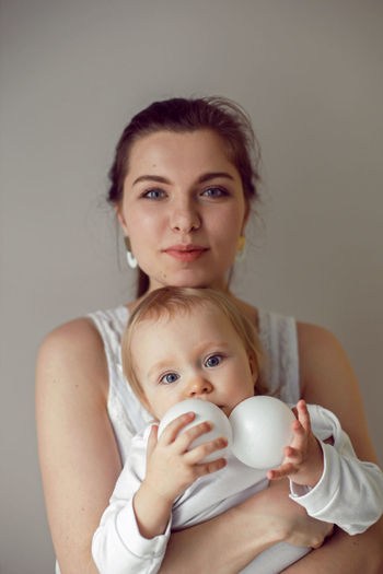 Mother holds her daughter a child in her arms stands next to the window in white clothes at home