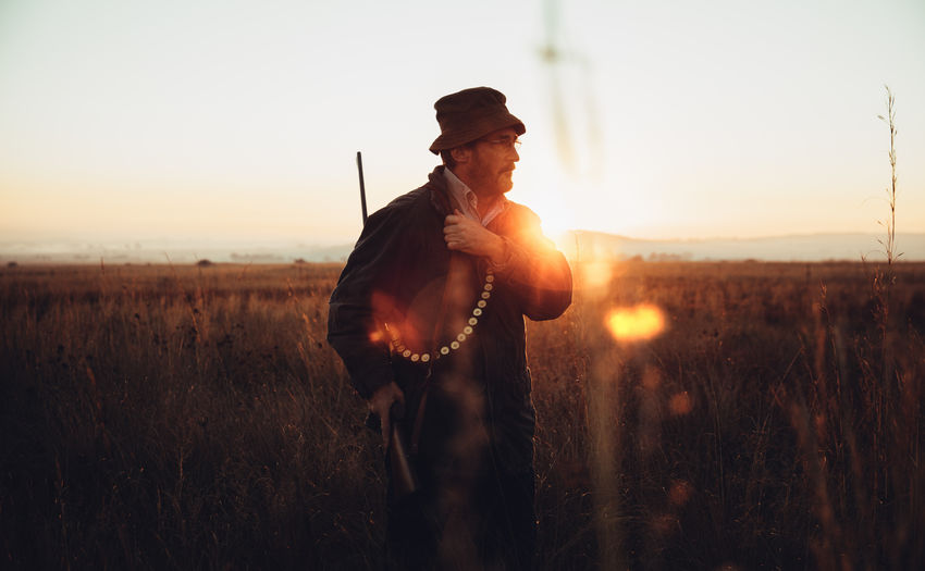 Hunter with shotgun and cartridge standing in field against sky