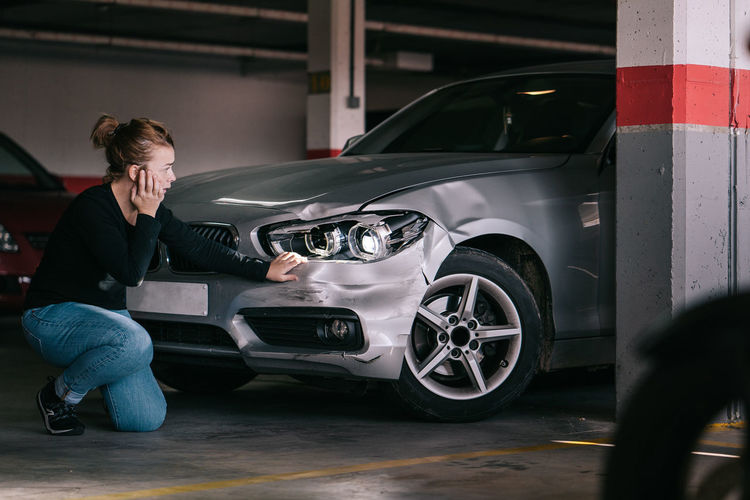 Side view of upset young lady in casual clothes sitting on knees and touching damaged car headlights after accident in parking space