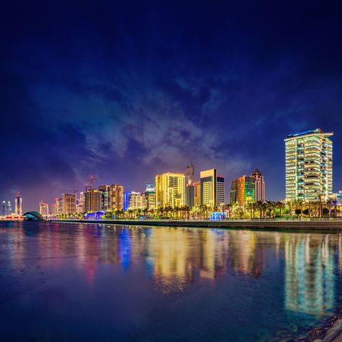 Illuminated buildings by river against blue sky at dusk