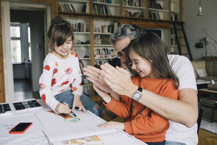 Happy father gesturing while helping daughter in doing homework by girl drawing at table
