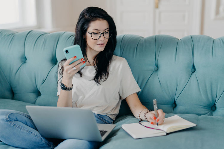 Young woman with mobile phone and laptop working on sofa