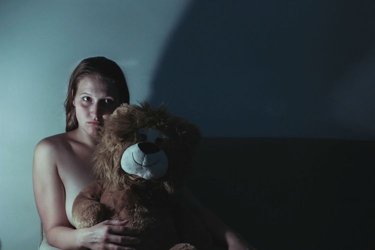 Portrait of naked woman holding teddy bear while sitting darkroom
