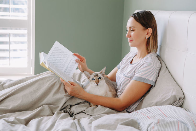 Woman reading book while sitting on bed with cat