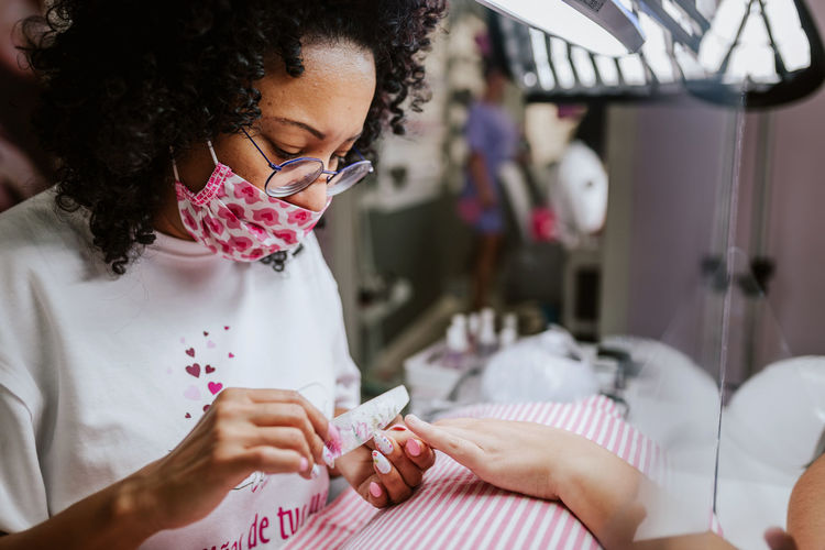 Side view of concentrated african american nail artist using file and polishing nails of crop customer during manicure in beauty studio