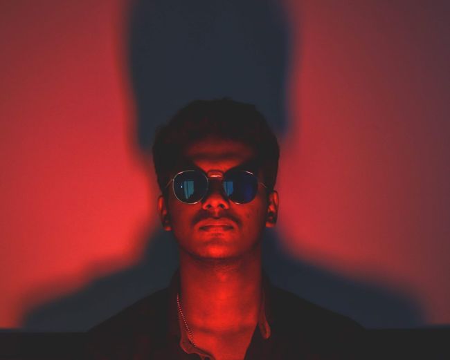 Portrait of young man wearing sunglasses in illuminated room against wall
