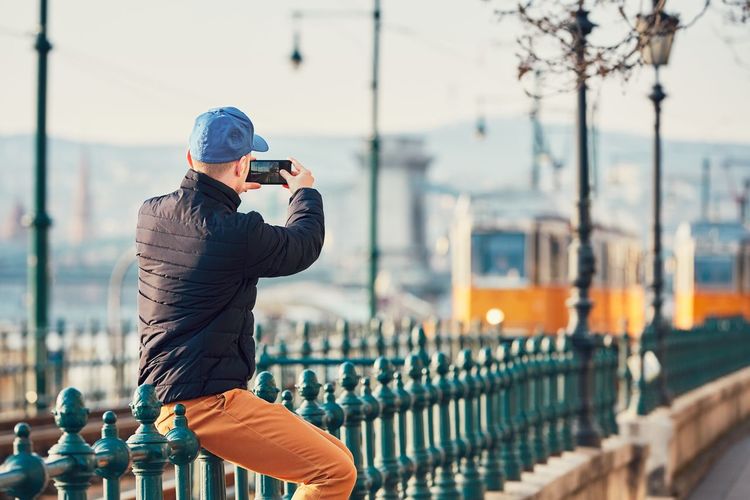 Rear view of man photographing while sitting on railing