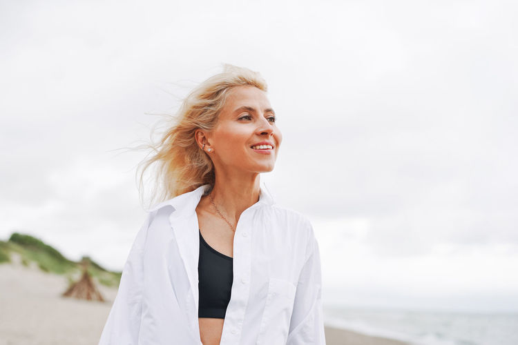 Portrait of elegant blonde woman in white shirt on sand beach at storm sea