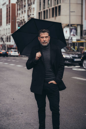 Pensive successful mature bearded male entrepreneur in black standing under umbrella during walk in street in rainy day