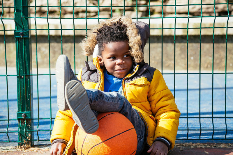 Smiling boy with basketball sitting by fence at court