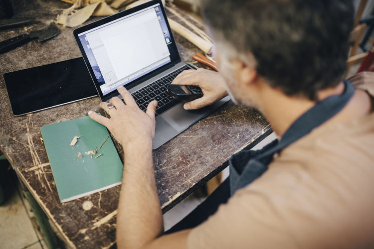 High angle view of male craftsperson using laptop at workbench in workshop