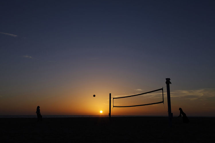 Silhouette people playing beach volleyball against sky during sunset