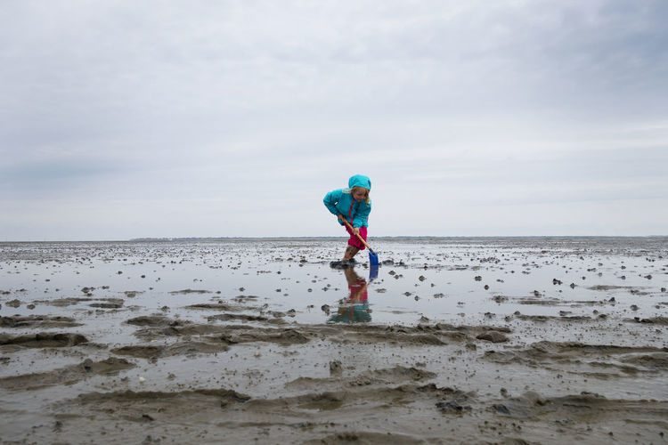 Girl playing in mud flat with shovel against sky at lower saxon wadden sea national park, germany