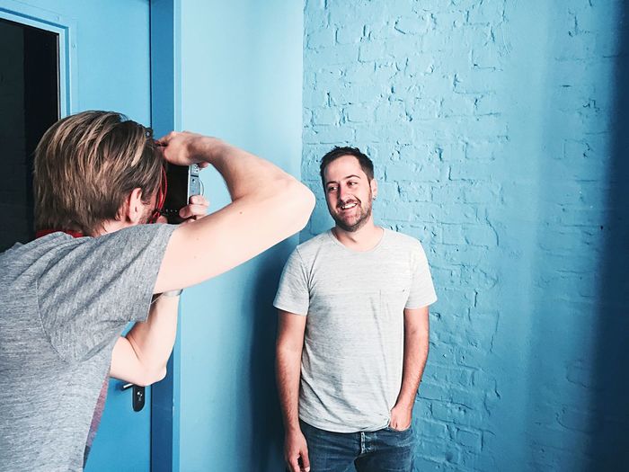 Man photographing friend standing by blue wall