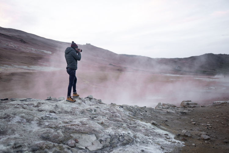 Full body side view of male traveler in outerwear standing on rocky ground and photographing steaming hot springs in iceland
