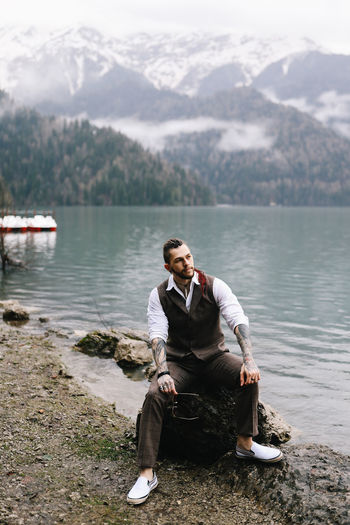 Young man sitting on lake against mountains