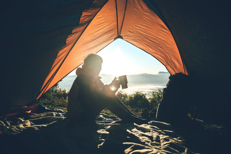 Man with mug in tent sitting on mountain against bright sun in sky during sunrise