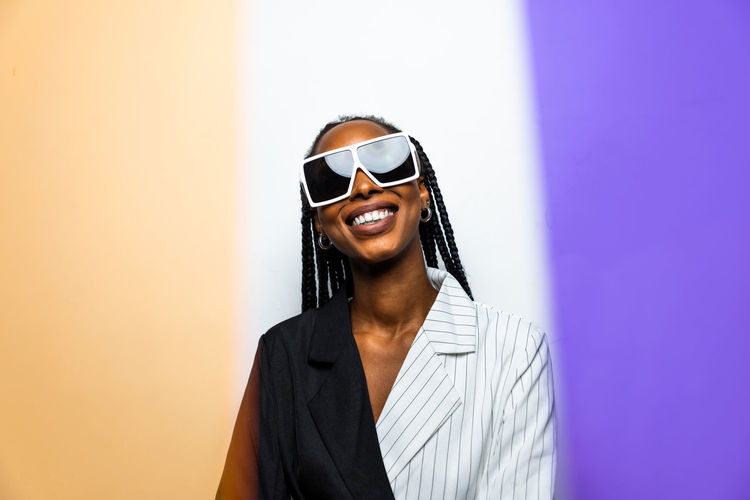 Portrait of smiling young woman wearing sunglasses standing against wall