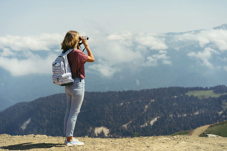 Young woman with a backpack in the mountains looks at the landscape through binoculars. 
