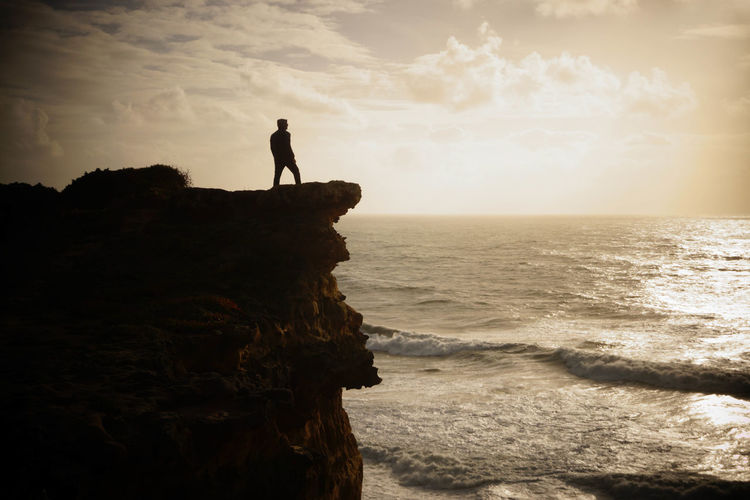 Silhouette man standing on cliff near sea against sky during sunset