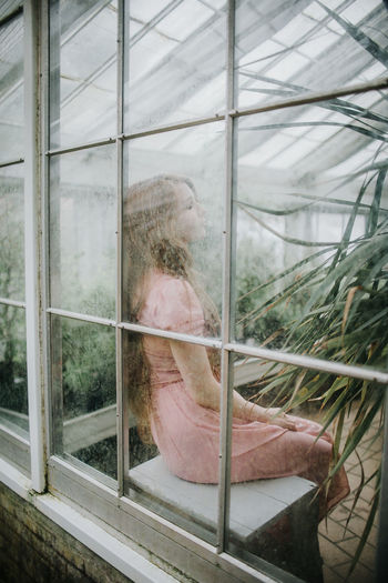 Through glass side view of young female in dress sitting in hothouse with green plants