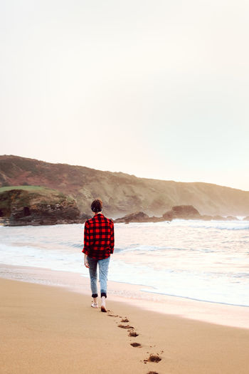 Back view of young short haired hipster female in checkered shirt and jeans walking on beach and leaving footprints on wet sand